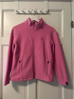 Rosa fleecejacka med dragkedja - Mysig rosa fleecejacka med hög krage och hel dragkedja framtill. Två fickor med dragkedja och broderad logga på bröstet. Perfekt för lager-på-lager, träning och chill dagar. Tillverkad i mjukt fleecematerial.🥰