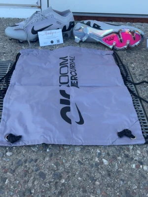 Nike Air Zoom Mercurial fotbollsskor - Säljer ett par grå Nike Air Zoom Mercurial fotbollsskor med svart swoosh och detaljer. Sulan har en cool metallic finish med rosa och silver, samt dobbar för bra grepp. Skorna är låga och kommer med en matchande grå påse med logga.