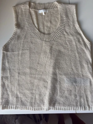 Beige stickad ärmlös topp - Säljer en beige stickad topp med bred ribbad kant vid halsen och ärmlös design. Toppen har en avslappnad passform och är glittrig.