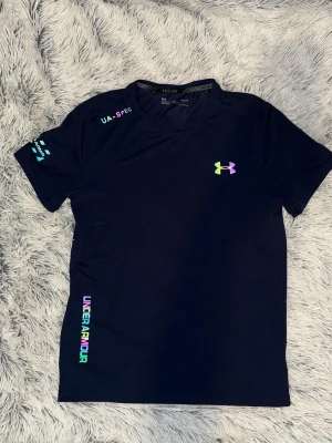 tshirt från Under Armour - Snygg svart tränings-tshirt från Under Armour med färgglada loggor och tryck på bröstet, axeln och sidan. Tillverkad i lätt och snabbtorkande funktionsmaterial, perfekt för löpning eller gym. Rund halsringning och korta ärmar för maximal rörelsefrihet.
