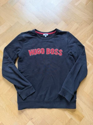 Svart sweatshirt från Hugo Boss - Cool svart sweatshirt från Hugo Boss med röd och vit broderad logga på bröstet. Klassisk rund halsringning och ribbade muddar vid ärmslut och nederkant. Perfekt för en avslappnad och stilren look.