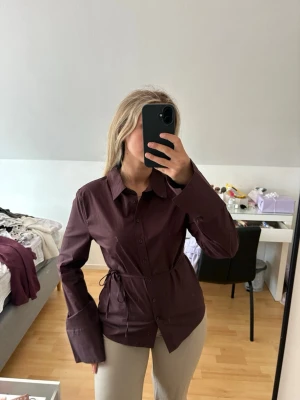 Mörkbrun skjorta med knyt  Shaped tie blouse - Snygg mörkbrun skjorta med knappar framtill och knyt i midjan för en mer markerad siluett. Skjortan har klassisk krage och långa ärmar med breda manschetter. Perfekt för dig som vill ha en stilren men ändå trendig look. Nypris 400kr. Prislapp kvar.