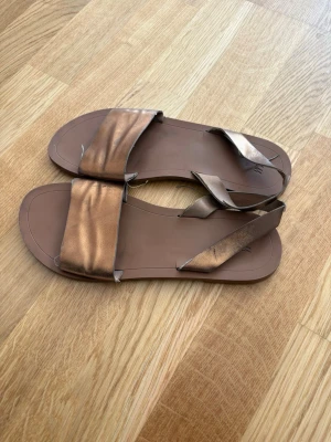 Bronsfärgade sandaler med bred rem - Säljer ett par stilrena sandaler i bronsfärgat material med bred rem över foten och smalare rem runt hälen. Sandalerna har en platt sula och minimalistisk design, perfekta för sommaren. 