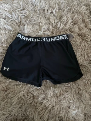 Svarta träningsshorts från Under Armour - Säljer ett par svarta träningsshorts från Under Armour med bred resår i midjan där loggan syns tydligt. Shortsen har en sportig look och är tillverkade i ett lätt och snabbtorkande syntetmaterial, perfekta för intensiva träningspass.