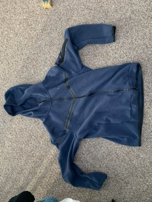 Mörkblå hoodie med dragkedja Nike - Snygg mörkblå hoodie med hel dragkedja framtill och svarta detaljer vid fickor och ärmar. Hoodien har en modern design med huva och praktiska dragkedjeförsedda fickor på både bröstet och ärmen. Perfekt för en avslappnad och sportig stil.
