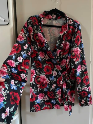 Blommig omlottblus med knytband - Snygg omlottblus från Gina Tricot med svart botten och färgglatt blommönster i rött, rosa och blått. Blusen har långa ärmar och ett knytband i midjan för en feminin siluett. Tillverkad i ett glansigt, mjukt material som känns lyxigt.