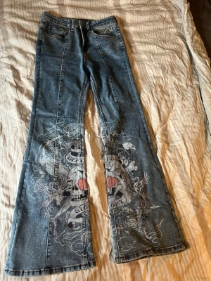Ed Hardy jeans med tryck - Unika blå Ed Hardy jeans med utsvängda ben och stort tryck med dödskallar, hjärtan och text på framsidan. Klassisk femficksmodell med knappgylf och tydliga kontrastsömmar. Perfekta för dig som vill sticka ut med cool streetstyle.