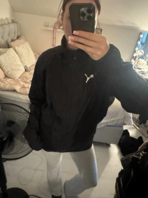 Svart vindjacka från Puma - Snygg svart vindjacka från Puma med dragkedja framtill samt två fickor med dragkedja. Tillverkad i lätt och vindtätt material. Finns även en luva som är invikt i kragen.