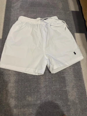 Vita badbyxor från Polo Ralph Lauren - Säljer ett par stilrena vita badbyxor från Polo Ralph Lauren med elastisk midja och dragsko. Klassisk passform, fickor i sidorna och en bakficka med lock. Liten mörkblå logga broderad framtill. Perfekta för stranden eller poolen.