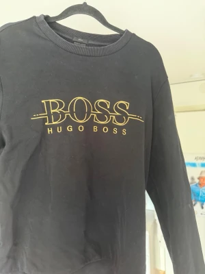 Svart sweatshirt från Hugo Boss - Svart/ mörkblå sweatshirt från Hugo Boss med gul logotyp och text på bröstet. Tröjan har rund halsringning, lång ärm och är i slim fit-modell. Använd 1 gång , skick 9/10.