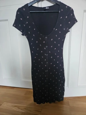 Svart blommig klänning Divided H&M - Svart kortklänning från Divided H&M med små blommiga mönster i vitt och rosa. Klänningen har korta ärmar, v-ringning och knappar framtill. Tillverkad i ribbad bomull för en bekväm och slim passform.
