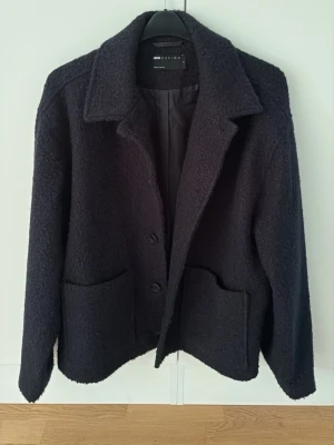 Svart cardiganjacka från ASOS DESIGN - Stilren svart cardiganjacka från ASOS DESIGN med två stora framfickor och klassisk krage. Jackan är tillverkad i ett mjukt, strukturerat material och har knäppning framtill. Perfekt för lager på lager och enkel att matcha med olika outfits.