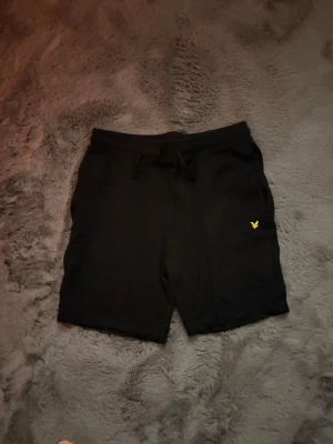 Svarta shorts från Lyle & Scott - Säljer ett par svarta shorts från Lyle & Scott med klassisk gul logga på vänster ben. Shortsen har snörning i midjan och är tillverkade i mjuk bomull, perfekta för en avslappnad stil.