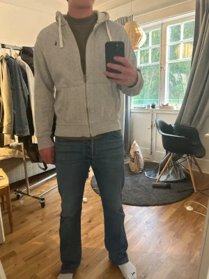 Grå hoodie från Polo Ralph Lauren - Säljer en grå zip-hoodie från Polo Ralph Lauren med huva och vita dragsnören. Klassisk broderad logga på bröstet och två fickor framtill. Tillverkad i mjukt sweatshirtmaterial som är perfekt för chill dagar. Enkel och stilren design som funkar till det mesta.
