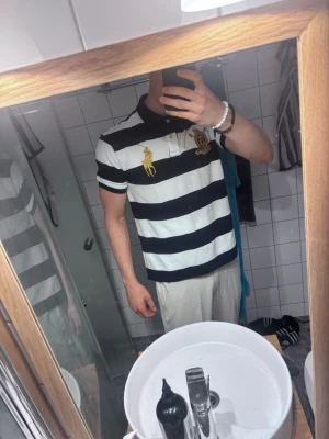 Randig pikétröja från Ralph Lauren Storlek L - Säljer en klassisk pikétröja från Ralph Lauren med breda svarta och vita ränder. Tröjan har korta ärmar, krage och knappar framtill samt broderad logga i gult och ett emblem på bröstet.