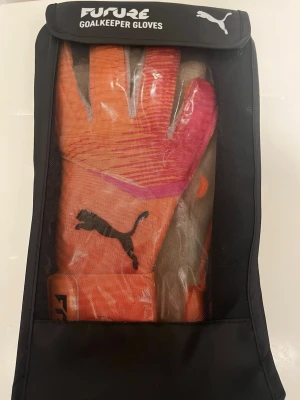 Puma Future målvaktshandskar orange - Säljer ett par grymma Puma Future målvaktshandskar i orange med rosa och lila detaljer. Handskarna har svart Puma-logga och bred kardborrestängning vid handleden. Perfekta för dig som vill sticka ut på planen och få bra grepp på bollen.