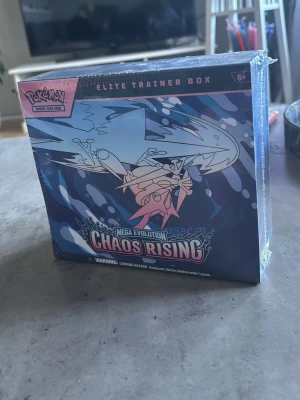 Pokémon Elite Trainer Box Chaos Rising - Säljer en Pokémon Elite Trainer Box Mega Evolution Chaos Rising. Boxen har ett blått och svart mönster med illustration av en drake och coola detaljer. Perfekt för dig som samlar eller spelar Pokémon Trading Card Game.