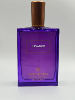 Lavande parfym från Molinard - En elegant parfymflaska i lila glas med guldlock från Molinard. Flaskan har en stilren, rektangulär form och texten 'Lavande' på framsidan. Doften är inspirerad av lavendel och flaskan har exklusiva guldiga detaljer.