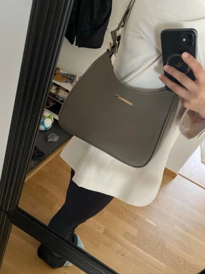 Olivgrön axelväska i konstläder - Snygg olivgrön axelväska i mjukt konstläder med minimalistisk design. Väskan har guldfärgade metalldetaljer, justerbar axelrem och dragkedja upptill. Perfekt storlek för det viktigaste och passar till många olika outfits.