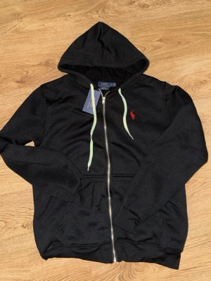Svart hoodie från Polo Ralph Lauren - Svart hoodie med dragkedja från Polo Ralph Lauren. Klassisk design med huva och vita dragsnören samt röd broderad logga på bröstet. Tillverkad i mjukt material och har fickor framtill. Perfekt för en avslappnad och stilren look. Storlek M. Aldrig använd