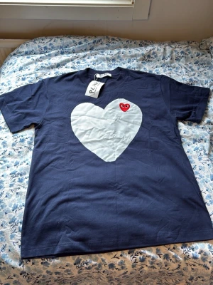 Blå Comme des Garçons PLAY t-shirt - Mörkblå t-shirt från Comme des Garçons PLAY med stort vitt hjärta och ett litet rött hjärta med ögon på bröstet. Klassisk rund hals och korta ärmar. Tillverkad i mjuk bomull, perfekt för en trendig och avslappnad stil. Helt ny aldrig använd storlek M