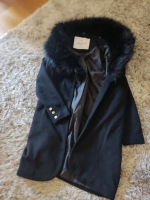 Svart kappa med fuskpäls från Chiara Forti - Stilren svart kappa från Chiara Forti Milano med stor krage i mjuk svart fuskpäls. Kappan har slät yta, knappar vid ärmsluten och helsvart foder. Perfekt för dig som vill ha en elegant och trendig look.