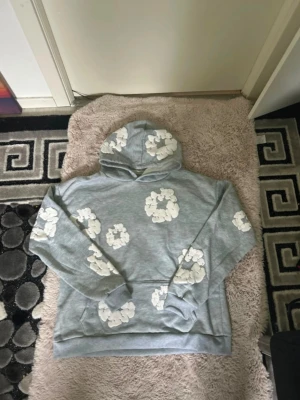 Denim tears hoodie - Snygg grå hoodie med vita blommönster över hela tröjan. Hoodien har en klassisk huva och magficka framtill. Perfekt för dig som gillar en avslappnad och trendig stil.