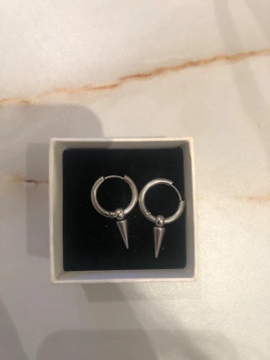 Silvriga hoops med spetsdetalj - Säljer ett par coola örhängen i form av silvriga hoops med spetsiga koner som hänger ner. De har en minimalistisk men edgy design och passar perfekt för dig som gillar att sticka ut. Materialet ser ut att vara metall i blank finish. Använd endast ett fåtal gånger😊