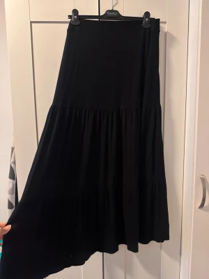 Svart långkjol i mjukt tyg - INDISKA - En svart långkjol med volangdetaljer och hög midja. Kjolen har en flowy passform och är tillverkad i ett mjukt, stretchigt material som känns bekvämt. Perfekt att styla med en crop top eller stickad tröja.