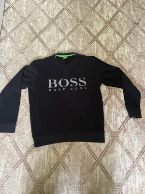 Svart sweatshirt från Hugo Boss - Säljer en svart sweatshirt från Hugo Boss med stort BOSS-tryck på bröstet. Tröjan har rund halsringning, långa ärmar och ribbade muddar. Perfekt för en clean och stilren look.