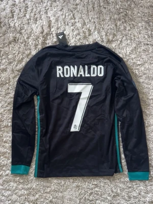 Real Madrid Ronaldo långärmad tröja - Snygg långärmad Real Madrid-tröja från Adidas med Ronaldo 7-tryck på ryggen. Svart med turkosa detaljer, klubbmärke och sponsorlogga framtill samt Champions League-märken på ärmarna. Tillverkad i lätt och ventilerande material.