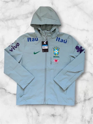 Nike Brasil vindjacka med huva - Ljusgrå vindjacka från Nike med Brasil-logga och sponsormärken som Itaú och Vivo. Jackan har hel dragkedja, huva och tryckta detaljer i blått, grönt och lila. Perfekt för dig som gillar sportig stil och vill sticka ut med landslagskänsla.