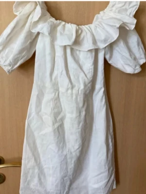 Sommarklänning  - Söt vit kortklänning med stora volanger längs urringningen och puffiga korta ärmar. Klänningen har en feminin och luftig känsla, perfekt för varma dagar. Tillverkad i ett lätt tyg som ger en avslappnad men ändå chic look.