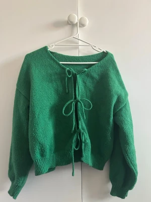 Grön stickad tröja med knytning - Cool grön stickad tröja med tre knytband framtill. Tröjan har en lös passform, långa ärmar och ribbade muddar. Perfekt för lager-på-lager och att styla med jeans eller kjol. Materialet känns mjukt och är troligtvis akryl eller ullblandning.