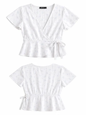 Vit omlottopp med knyt och blommönster - Söt vit omlottopp med ljusblått blommönster och knyt i sidan. Toppen har korta ärmar, v-ringning och volang nedtill för en feminin look. Perfekt till jeans eller kjol för en fräsch sommarstil.