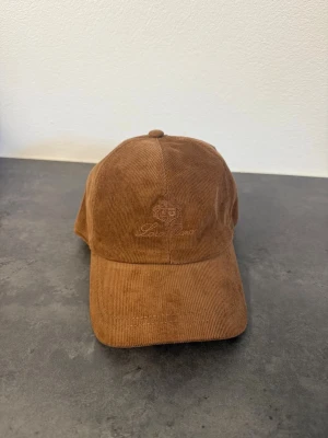 Brun keps i manchester från Loro Piana - Snygg brun keps i manchestertyg från Loro Piana med broderad logga framtill. Klassisk böjd skärm och justerbar baksida. Perfekt accessoar för en avslappnad och trendig look.