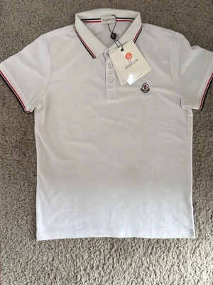 Vit Moncler pikétröja med röd detalj - Klassisk vit pikétröja från Moncler med korta ärmar och krage. Tröjan har röda och svarta ränder på krage och ärmslut samt Moncler-logga på bröstet. Perfekt till sommaren och tillverkad i mjuk bomull för en skön känsla.