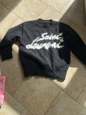 Svart Saint Laurent stickad tröja - Cool svart stickad tröja från Saint Laurent med vit fuzzy logga framtill. Tröjan har rund halsringning, ribbade muddar och är tillverkad i Italien. Perfekt för dig som gillar streetstyle och exklusiva märken.