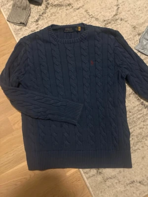 Marinblå kabelstickad tröja Polo - Snygg marinblå kabelstickad tröja från Polo Ralph Lauren med rund halsringning och diskret röd logga broderad på bröstet. Tröjan har ribbade muddar och är perfekt för lager-på-lager-stil. Klassisk och tidlös design.