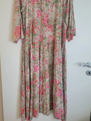 Blommig beige/rosa långklänning från Lindex  - Långklänning i beige med färgglatt blommigt mönster i rosa, orange och grönt. Klänningen har trekvartsärm och en lös, flowy passform som ger en avslappnad och romantisk känsla. Tillverkad i mjuk Viscose, perfekt för varma dagar.