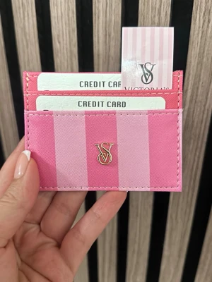 Rosa kortfodral från Victoria's Secret - Snyggt kortfodral i rosa och ljusrosa ränder från Victoria's Secret. Har tre fack för kort och ett guldfärgat VS-emblem på framsidan. Tillverkat i ett tåligt material med stilren design, perfekt för att hålla koll på dina kort.
