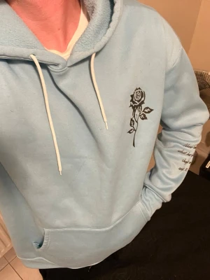 Ljusblå hoodie med ros-tryck - Ljusblå hoodie med vit dragsko och stor ficka framtill. På bröstet finns ett svart tryck av en ros och på ena ärmen står ett citat i svart text. Hoodien har en klassisk huva och är gjord i mjukt material, perfekt för en avslappnad stil.