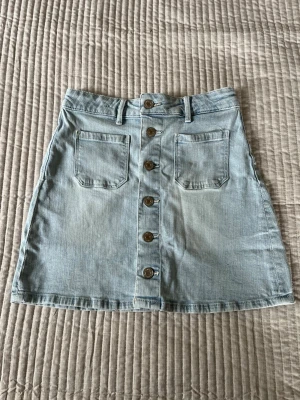Ljusblå jeanskjol med knappar - Snygg ljusblå jeanskjol med knappar framtill och snygga fickor som en extra detalj. Även hällor om man skulle vilja ha ett bälte. Går lätt att stayla till det mesta. Perfekt till sommarens alla tillfällen. 