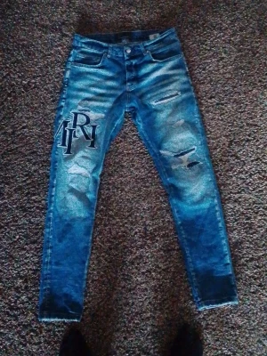 Amiri jeans MX1 size30 Blå slitna jeans med tryck - Säljer ett par blå jeans med slitningar och tryckta bokstäver på både fram- och baksida. Jeansen har smal passform, klassiska fem fickor och är gjorda i denim. Snygga detaljer med slitna partier och stora bokstäver på låret och vaden.