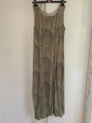 Långklänning med zigzagmönster - Säljer en ärmlös långklänning med svart och beige zigzagmönster. Klänningen har rund halsringning och ett mjukt, följsamt tyg som ger en avslappnad siluett. Perfekt för dig som gillar grafiska mönster och vill ha en unik look.