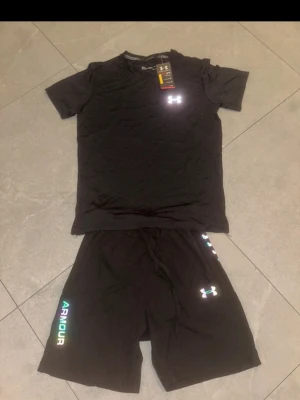 Svart träningsset från Under Armour - Snyggt svart träningsset från Under Armour med t-shirt och shorts. Båda plaggen har logga framtill och shortsen har texten 'ARMOUR' i färg längs benet. Perfekt för gymmet eller löpning. Materialet är lätt och andas, vilket gör det bekvämt under träning.