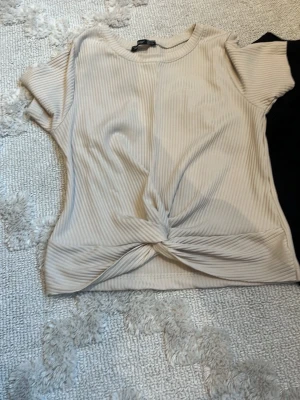 Två ribbade toppar med knutdetalj - Säljer två snygga ribbade toppar i beige och svart. Båda har korta ärmar, rund halsringning och en trendig knutdetalj framtill. Perfekta att styla till jeans eller kjol för en cool look.