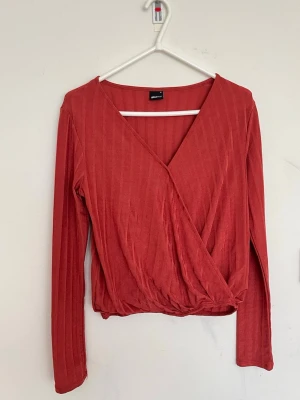 Röd omlottopp med ribbad struktur - Trendig röd omlottopp från Gina Tricot med ribbad struktur och långa ärmar. Toppen har en snygg v-ringning och croppad passform som ger en modern vibe. Perfekt att styla med höga jeans eller kjol.