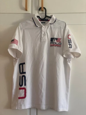 Vit Polo USA Sailing Team piké - Vit pikétröja från Polo med USA-flagga på ärmen och stora tryck med texten 'USA' och 'Sailing Team' fram och bak. Marinblå och röda detaljer, klassisk krage med knappar och korta ärmar. Perfekt för dig som gillar sportig stil.