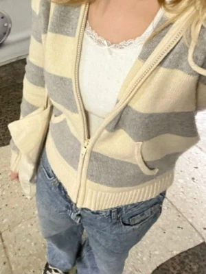 Randig hoodie med dragkedja - Säljer en mysig hoodie från Brandy Melville med breda beige och ljusblå ränder. Hoodien har dragkedja framtill, två fickor och en huva med snören. Tillverkad i mjukt stickat material, perfekt för chill dagar. Passar till jeans eller mjukisbyxor.